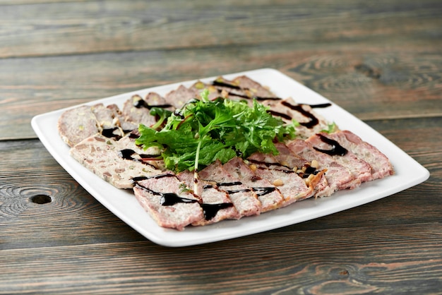 Primer plano de un plato cuadrado blanco servido con rebanadas de carne rellenas, decorado con hojas verdes y salsa de soja sobre la mesa de madera.