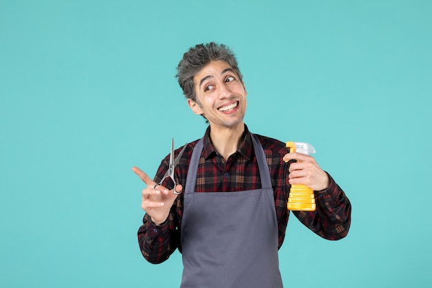 Primer plano de preguntando peluquero masculino vistiendo delantal gris y sosteniendo una botella de spray de tijeras sobre fondo de color azul suave