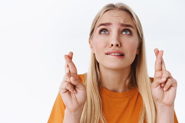 Primer plano preocupado muy nervioso mujer caucásica rubia en camiseta naranja con los dedos cruzados espero que Dios escuche oraciones como cejas de surco intenso ansiosamente suplicando soporte fondo blanco