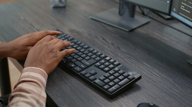 Primer plano de programador afroamericano manos escribiendo código en el teclado mientras mira pantallas de computadora con interfaz de programación. Ingeniero de sistemas sentado en el escritorio desarrollando algoritmos.