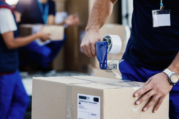 Primer plano de un repartidor cerrando la caja de cartón con una cinta mientras prepara los paquetes para su envío