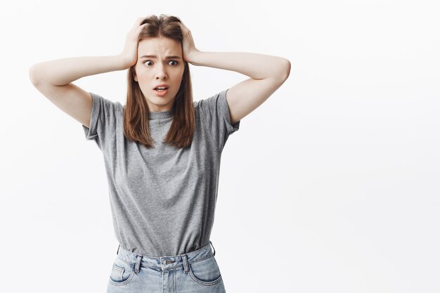 Primer plano retrato de atractiva joven caucásica con cabello oscuro en ropa casual mirando a un lado con expresión asustada, apretando la cabeza con las manos, teniendo pánico después de perder sus llaves.