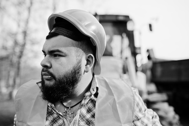 Primer plano retrato de barba brutal trabajador hombre traje trabajador de la construcción en casco naranja de seguridad contra traktor