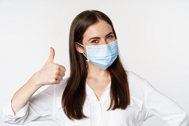 Foto gratuita primer plano retrato, cara de mujer con máscara médica sonriendo, mostrando los pulgares hacia arriba, de pie sobre fondo blanco