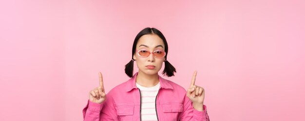 Primer plano retrato de una chica asiática con estilo que parece escéptica apuntando con el dedo hacia arriba con la cara sin gracia de pie sobre fondo rosa Copiar espacio