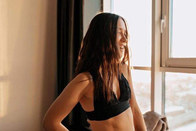 Primer plano retrato de una chica bastante encantadora con el pelo ondulado vestido con la parte superior deportiva mirando a la ventana a la luz del sol Yoga matutino en casa