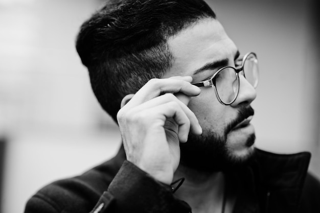Primer plano retrato de empresario de barba del Medio Oriente usar abrigo negro y anteojos de camisa azul contra el edificio de oficinas