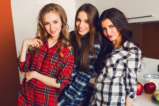 Primer plano retrato de estilo de vida interior de tres divertidos jóvenes amigos divertirse y fingir caras en la cocina. Ambiente de fiesta en casa. El uso de camisa a cuadros. Foto cálida y suave.