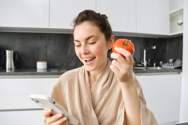 Foto gratuita primer plano retrato de una hermosa joven en la cocina sosteniendo tomate y teléfono inteligente compra productos frescos