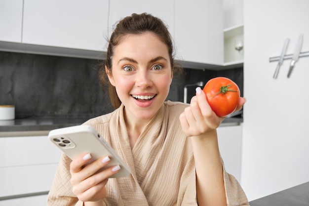 Foto gratuita primer plano retrato de una hermosa joven en la cocina sosteniendo tomate y teléfono inteligente compra productos frescos