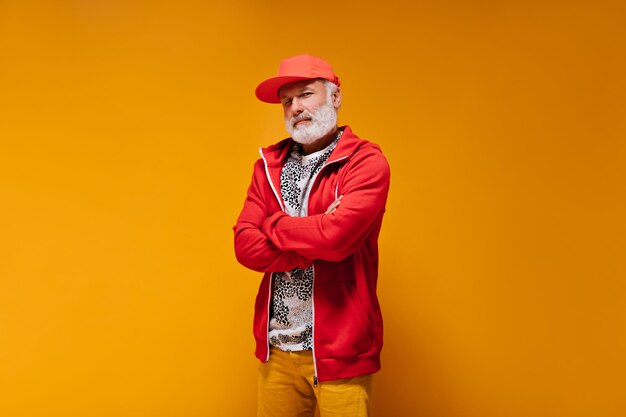 Primer plano retrato de hombre adulto en traje rojo sobre fondo naranja Chico guapo genial con barba gris en gorra y sudadera brillante posando