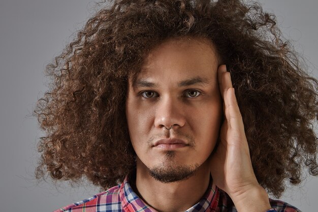 Primer plano retrato de hombre joven y guapo de piel oscura con cabello voluminoso, ojos marrones, mejillas regordetas y barba recortada posando aislada con mirada seria, sosteniendo la mano en su rostro