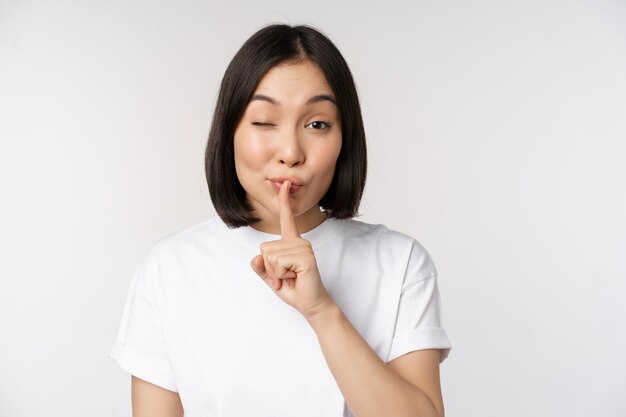 Primer plano retrato de joven hermosa chica asiática shushing tiene secreto mantener silencio silencio gesto presione el dedo a los labios de pie en camiseta sobre fondo blanco