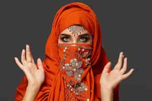 Foto gratuita primer plano retrato de una linda mujer joven con hermosos ojos ahumados y joyas finas en la frente, usando el hiyab de terracota decorado con lentejuelas. ella está mirando asustada en un fondo oscuro. tararear