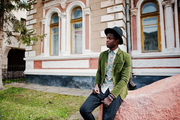 Primer plano retrato de moda de hombre afroamericano negro con chaqueta de terciopelo verde y sombrero negro sentado en la antigua mansión de fondo de escaleras