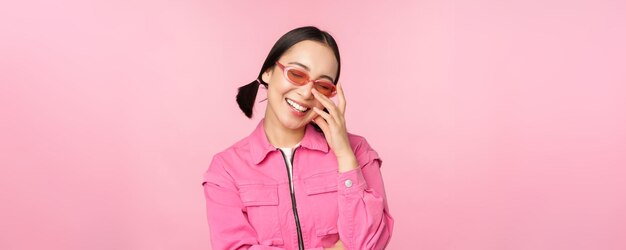 Primer plano retrato de mujer asiática con estilo en gafas de sol sonriendo mirando halagado riendo coqueto de pie sobre fondo rosa