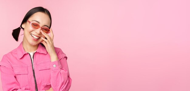 Primer plano retrato de mujer asiática con estilo en gafas de sol sonriendo mirando halagado riendo coqueto de pie sobre fondo rosa