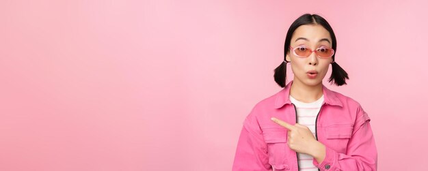 Primer plano retrato de una mujer asiática jadeando sorprendida señalando con el dedo el anuncio de pancarta de pie sobre fondo rosa