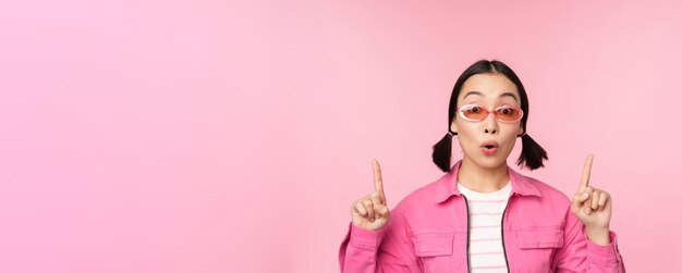 Primer plano retrato de una mujer asiática jadeando sorprendida señalando con el dedo el anuncio de pancarta de pie sobre fondo rosa