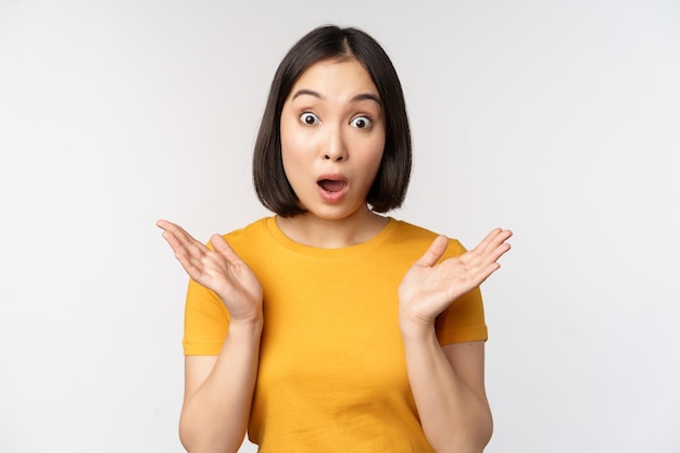 Primer plano retrato de una mujer asiática que parece sorprendida cara de asombro mirando impresionada a la cámara de pie sobre fondo blanco en camiseta amarilla