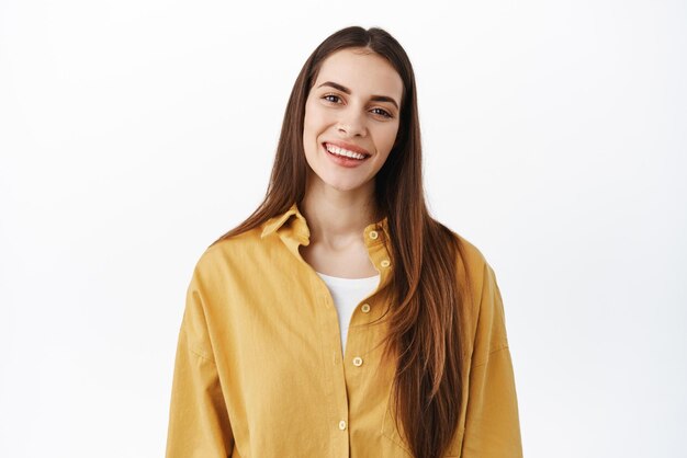 Primer plano retrato de mujer caucásica feliz con cabello largo natural maquillaje desnudo sonriendo e inclinando la cabeza luciendo amistoso y positivo de pie sobre fondo blanco