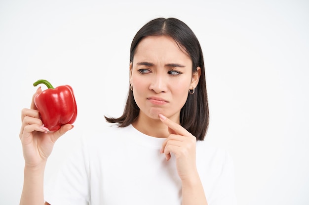 Foto gratuita primer plano retrato de mujer coreana pensativa mira pimiento rojo con cara de pensamiento aislado en blanco b