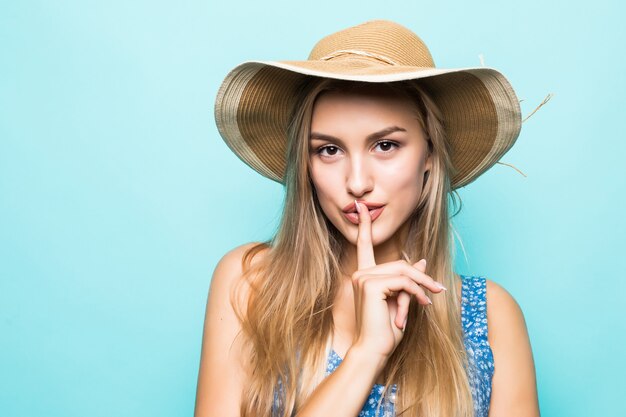 Primer plano retrato de mujer encantadora europea vistiendo un gran sombrero de paja mostrando el dedo en los labios para mantener el secreto aislado sobre fondo azul.