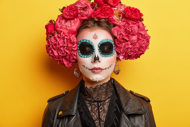 Primer plano retrato de mujer joven demuestra arte facial, usa maquillaje profesional, corona y chaqueta se prepara para la fiesta de disfraces de Halloween, mantiene los ojos cerrados