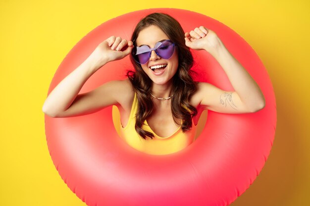 Primer plano retrato de mujer joven con estilo, chica con anillo de natación y gafas de sol de verano, lista para vacaciones en la playa, vacaciones, de pie sobre fondo amarillo.