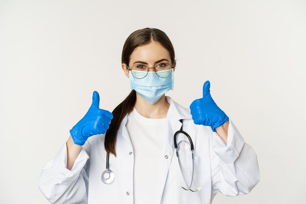 Primer plano retrato de mujer médico médico en máscara facial de coronavirus mostrando los pulgares hacia arriba y sonríe ...