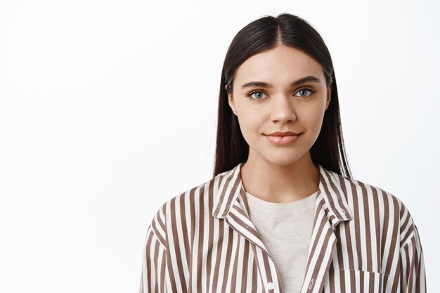 Primer plano retrato de mujer moderna y elegante en chaqueta de moda, con maquillaje de luz natural, sonriendo tiernamente y mirando al frente, de pie sobre la pared blanca