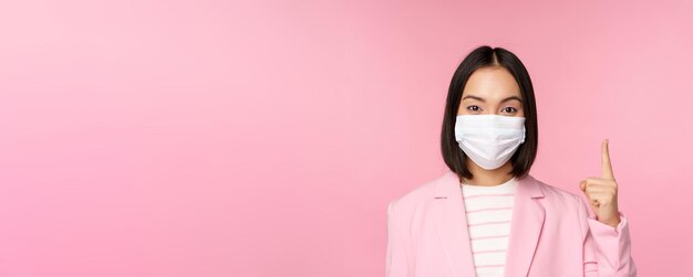 Primer plano retrato de mujer de negocios asiática con mascarilla médica y traje señalando con el dedo hacia arriba mostrando un anuncio