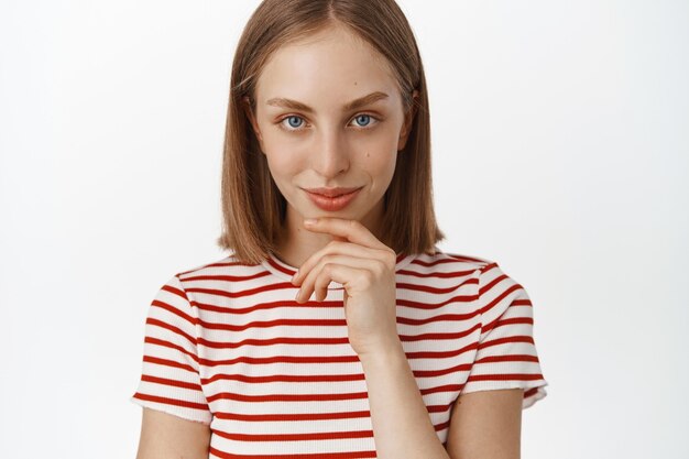 Primer plano retrato de mujer rubia con rostro limpio y natural sin maquillaje, mirando pensativo y decidido, sonriendo y mirando como si tuviera una idea, gran plan, de pie contra la pared blanca