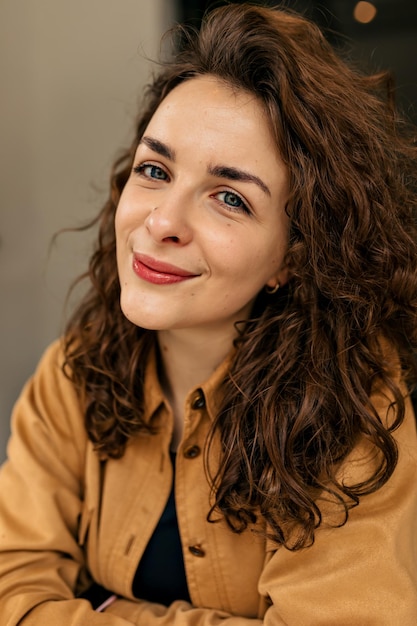 Primer plano retrato de tierna mujer encantadora con cabello rizado y maquillaje desnudo posando en la cámara con una sonrisa encantadora Mujer morena con camisa marrón se siente bien el fin de semana Concepto de confort en el hogar