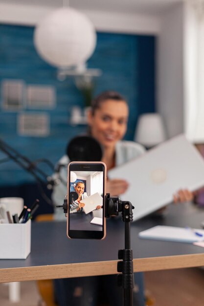 Primer plano de la revisión del producto de grabación de teléfonos inteligentes. Presentador de mujer de filmación de teléfono móvil haciendo pruebas de vlog de computadora portátil. Influenciador de las redes sociales haciendo recomendaciones de gadgets frente a la cámara.