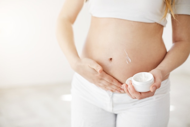 Primer plano de una rubia embarazada precoz cuidando la piel de su vientre con crema de vitamina.