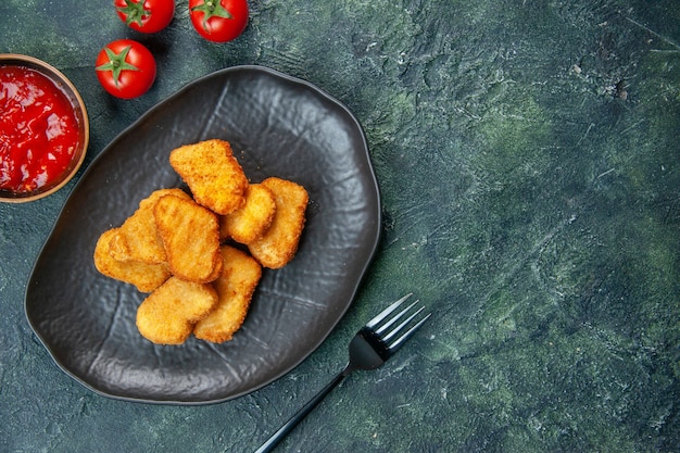 Primer plano de sabrosos nuggets de pollo en placa negra, tenedor de tomates en el lado derecho sobre una superficie oscura con espacio libre