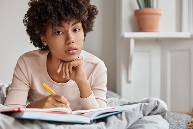 Primer plano de una seria estudiante universitaria de piel oscura hace tareas domésticas en la cama, escribe en un cuaderno con lápiz, mantiene la mano debajo de la barbilla