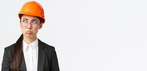 Primer plano de una sombría ingeniera asiática disgustada con casco de seguridad y traje de negocios que se queja de que se ve decepcionada y se queja con la cara de mal humor que no quiere trabajar los fines de semana y se ve molesta