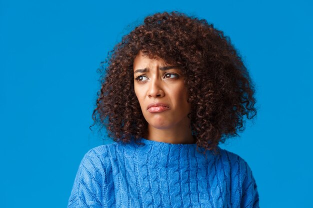 Primer plano sombrío y molesto, enfurruñada niña afroamericana de mal humor odia el clima frío nevado y la temporada de invierno, haciendo muecas mirando afuera disgustado, haciendo pucheros quejándose, pared azul.