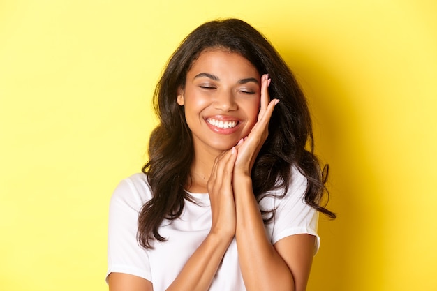 Primer plano de soñadora, hermosa mujer afroamericana, tocando suavemente la cara y sonriendo complacido, de pie contra el fondo amarillo