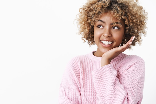 Primer plano de soñadora tierna y linda mujer afroamericana con estilo en suéter de invierno cálido tocando la mejilla y sonriendo encantada en la esquina superior izquierda disfrutando de una condición de piel perfecta