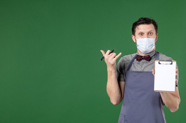 Primer plano de sonriente camarero masculino en uniforme con máscara médica y sosteniendo el libro de pedidos sobre fondo verde