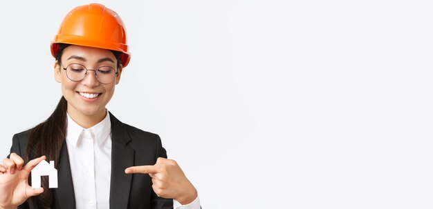 Primer plano de una sonriente ingeniera arquitecta asiática con casco y traje de negocios sosteniendo una miniatura casera y señalando con el dedo a una pequeña casa trabajando sobre la construcción de edificios de fondo blanco