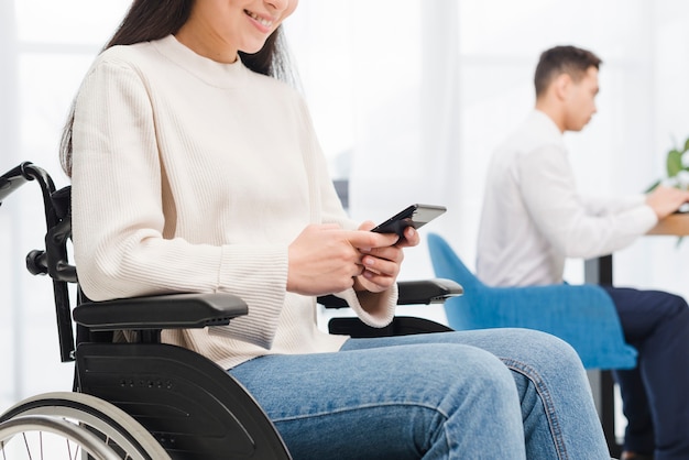 Primer plano de una sonriente joven discapacitada sentada en silla de ruedas usando un teléfono móvil frente a su colega masculino