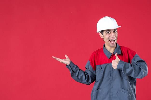 Primer plano de sonriente joven trabajador en uniforme con casco apuntando algo haciendo gesto ok en pared roja aislada