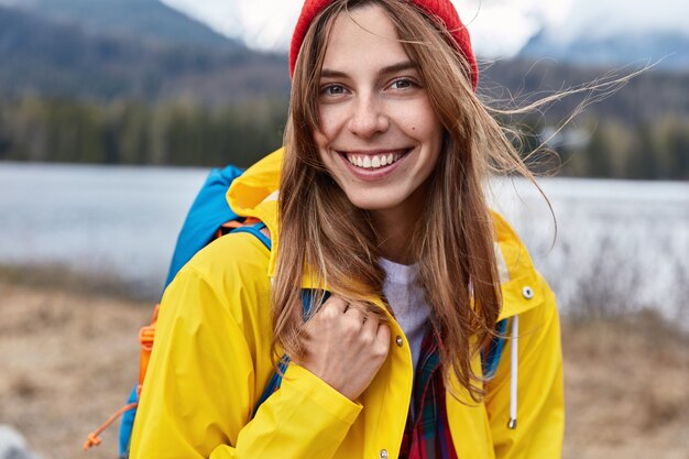 Primer plano de sonriente turista atractiva en abrigo amarillo, lleva mochila