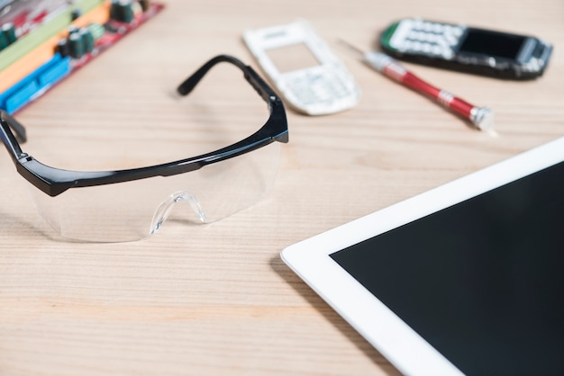 Primer plano de la tableta digital; Gafas de seguridad y teléfono móvil roto sobre fondo de madera