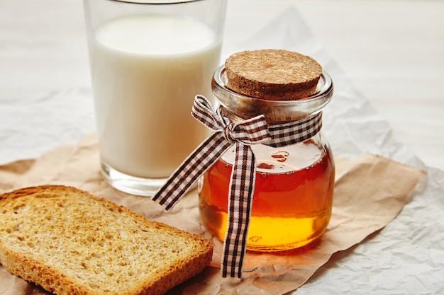 Primer plano de un tarro de miel con cinta bonita como regalo. Vaso desenfocado de leche y pan tostado de centeno seco alrededor. Todo en papel artesanal.