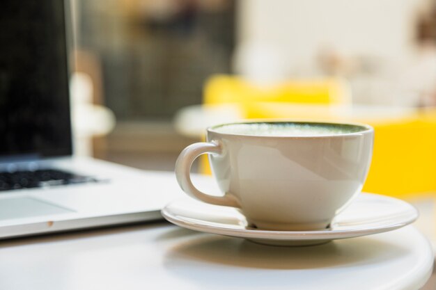 Primer plano de la taza blanca con café con leche verde cerca de la computadora portátil en la mesa blanca